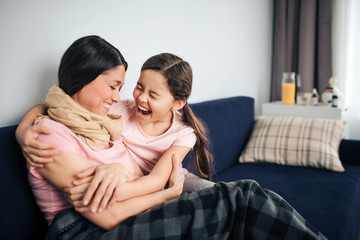 Happy and positive young woman sit together with her daughter on couch in room. They laugh out loud. Young woman is sick. Her throat is wrapped with scarf. Girl embrace mother.