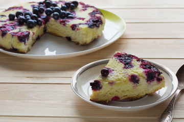 Slice of cake and baked round black currant cake on white plate on light wooden background. Shallow focus. Soft light