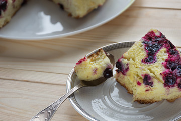 Slice of cake and baked round black currant cake on white plate on light wooden background. Shallow focus. Soft light