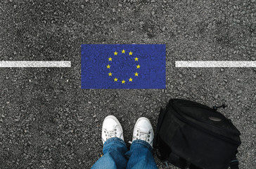 a man with a shoes and backpack is standing on asphalt next to flag of European Union and border