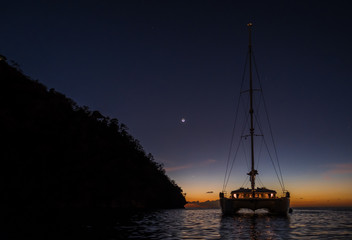 Dark night view on sailing boat anchored on open sea with black silhouette of island