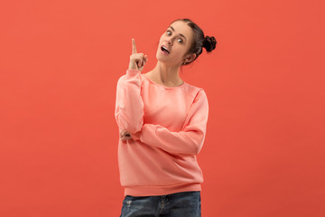 Happy woman standing and smiling isolated on coral studio background. Beautiful female half-length portrait. Young emotional woman pointing up. The human emotions, facial expression concept.