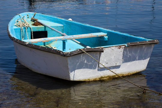 Closeup View of Dinghy Paddle Boat on Clear Water. Oar, rusty Chain and Rope Resting inside the Blue Watercraft. Vessel Looking Old and weathered from use