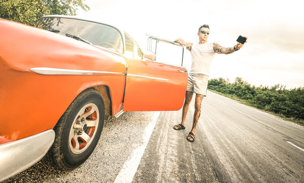 Young hipster fashion man with tattoo taking selfie with vintage car during road trip in Cuba - Travel wanderlust concept always connected on social media influencer lifestyle - Bright retro filter