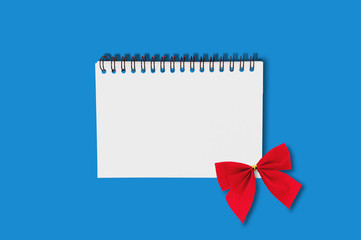 One paper notebook with spiral wire and red festive textile tied bow in center on blue table.  Top view. Holidays concept
