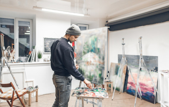 Portrait of an artist's student, drawing a painting in a studio. Young painter paints a new oil painting in his own cozy studio. Creative work.