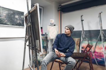 Fototapeta na wymiar Portrait of a professional artist in the interior of a cozy home studio. Painter sits on a chair near the easel with the host and palette, looking at a serious look at the picture. Painting Concept.