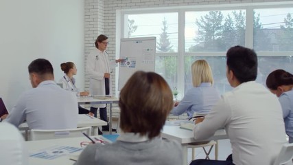 Experienced male doctor in lab coat explaining something on flipchart and giving presentation to healthcare workers in seminar room while his female assistant with stethoscope writing down at desk