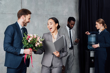 businessman presenting flowers to cheerful asian businesswoman with colleagues on background in waiting hall