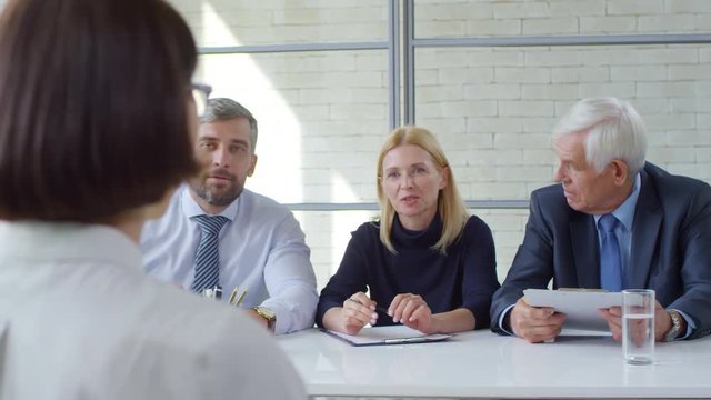 Tracking shot of bearded businessman, blond businesswoman and elderly manager in suit sitting at table and having conversation with female candidate during panel job interview