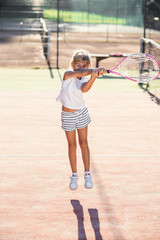 Female little tennis player in white sport uniform practice in hitting with tennis racket at the training on outdoor court on the sunset background.