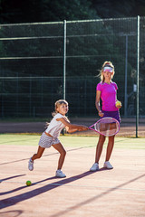 Obraz premium Professional young coach throws ball and little tennis player hits ball by racket during training on the outdoor court. Sport, health care concept.