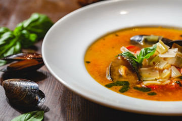 Soup with mussels and vegetables in white plate on wooden table. Close up
