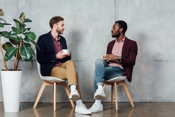 multiethnic men in casual clothes sitting, drinking coffee and talking in waiting hall