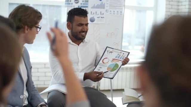 Cheerful middle eastern businessman showing financial diagram to colleagues and explaining it while sitting in circle in the office and discussing ideas