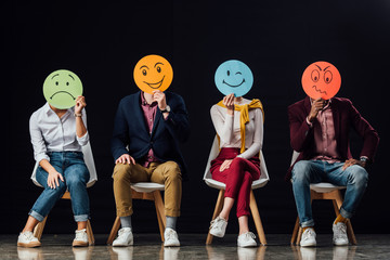 group of people sitting on chairs and holding face cards with various emotions isolated on black