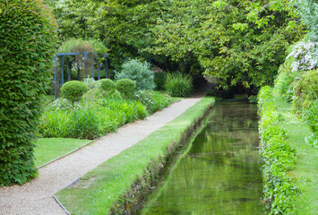 Water canal by a gravel walking path in a landscaped english garden, with mature trees, topiary shaped shrubs, on a summer day .