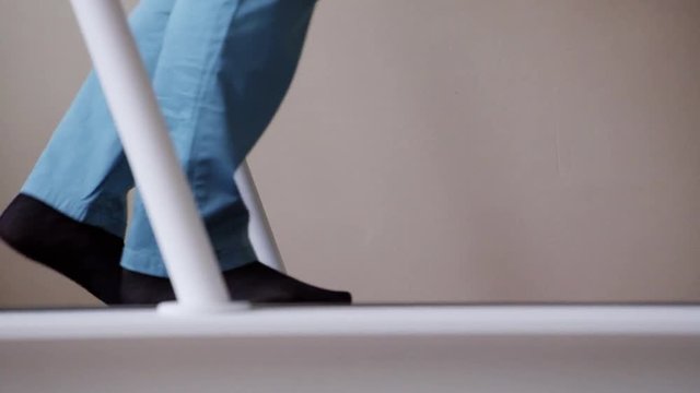 View of metallic medical device track with person's legs wearing blue pants and black socks slowly walking on it in room with white walls.