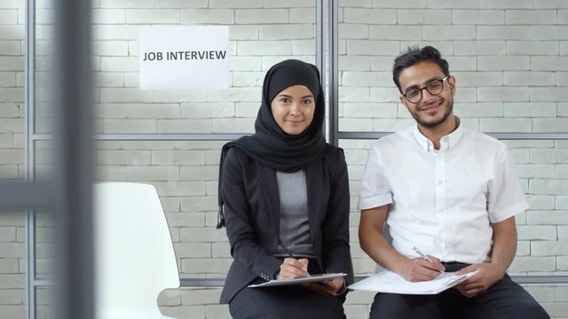 Tracking shot of cheerful young Muslim woman in hijab and smiling Arab man in glasses sitting in chairs in office reception area and smiling for camera while waiting for job interview