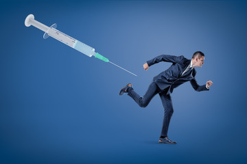 Fototapeta na wymiar A side view of a businessman running from a huge syringe floating in the air on a blue background.