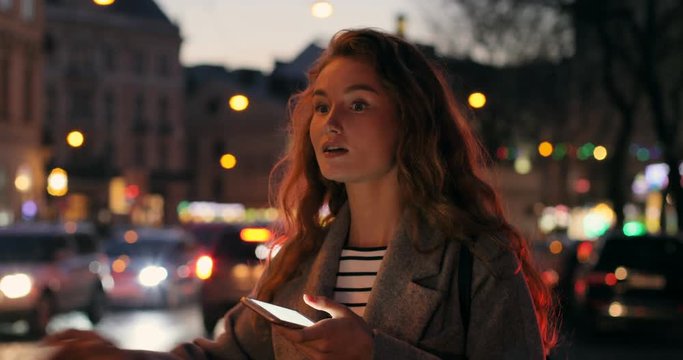 Close up of the smiled pretty young red haired woman typing and texting on the smartphone and then catching a taxi in the city street in the evening.