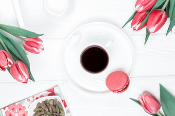 Black coffee in a white cup, cardamom, red tulips and raspberry macaron or macaroon on a wooden background. Top view