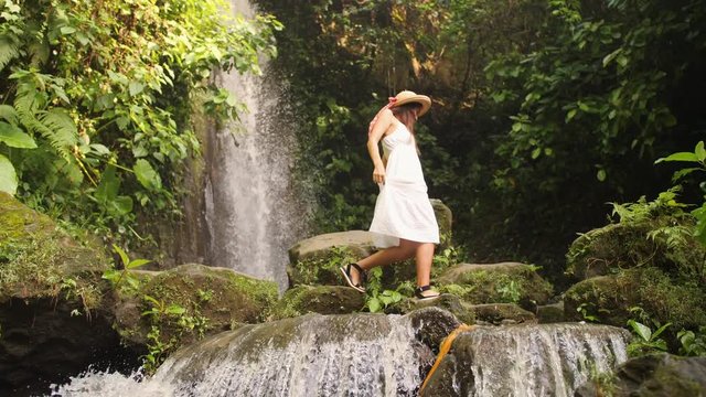 Attractive Girl in White Dress and Straw Hat Walking Barefoot near Small Waterfall in Tropical Rainforest Jungle. Carefree Lifestyle Travel 4K Slowmotion Footage. Bali, Indonesia.