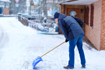 Man with snow shovel cleans sidewalks in winter during snowfall. Winter time in Europe. Young man in warm winter clothes. Snow and weather chaos in Germany. Snowstorm and heavy snowing. Schneechaos