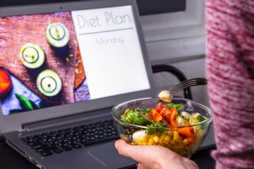 Bowl of vegetable salad in female hands near the laptop on the desktop. On the screen - planning a diet for weight loss from Monday