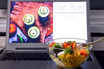 Bowl of vegetable salad near the laptop on the desktop. On the screen - planning a diet for weight loss from Monday