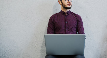Young pensive guy using laptop, wearing glasses on background of white.