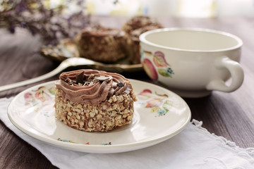 Biscuit cake sprinkled with nut crumb and decorated with chocolate cream on a white ceramic plate on a white napkin next to cup, closeup