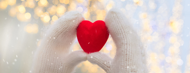 Female hands in white knitted mittens with red heart on glittering holiday background. St....