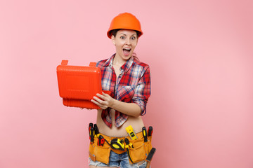 Strong young handyman woman in orange helmet, plaid shirt, denim shorts, kit tools belt full of instruments, toolbox isolated on pink background. Female in male work. Renovation and occupation concept