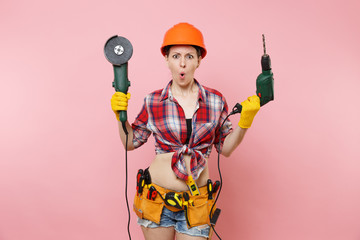 Energy fun handyman woman in gloves, protective orange helmet, kit tools belt full of instruments holding power saw electric drill isolated on pink background. Female in male work. Renovation concept