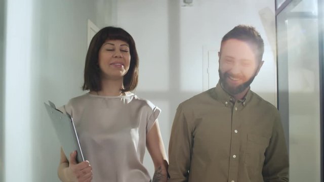 Dolly shot of cheerful businesswoman with document folder and smiling bearded businessman walking along office corridor and chatting