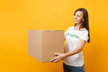Portrait of smiling woman in white t-shirt with written inscription green title volunteer with big cardboard box isolated on yellow background. Voluntary free assistance help, charity grace concept.