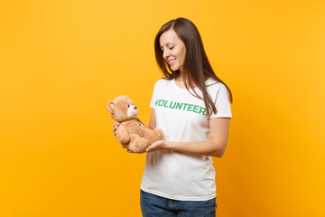 Portrait of woman in white t-shirt with written inscription green title volunteer hold teddy bear plush toy isolated on yellow background. Voluntary free assistance help, charity grace work concept.