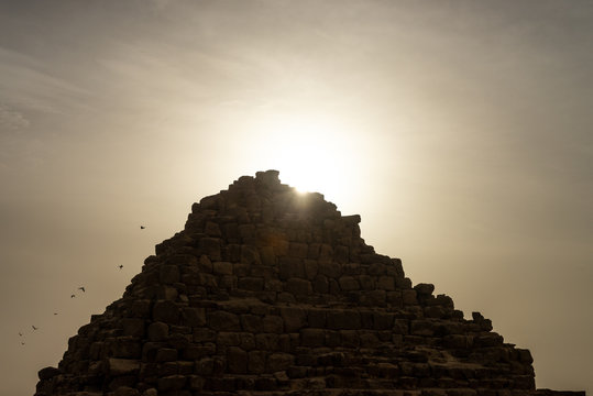 The Tomb of Queen Hetepheres in the Giza pyramid complex with star beams of the rising sun behind it and pigeons flying away.