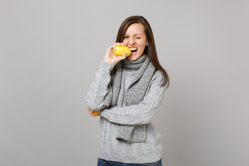 Cheerful young woman in gray sweater, scarf blinking, biting lemon isolated on grey background in studio. Healthy fashion lifestyle, people sincere emotions, cold season concept. Mock up copy space.