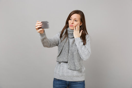 Sad young woman in sweater, scarf putting hand on cheek, doing selfie shot on mobile phone making video call isolated on grey background. Healthy fashion lifestyle people emotions cold season concept.