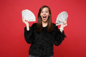 Surprised young woman in fur sweater with opened mouth holding fan of money in dollar banknotes, cash money isolated on red background. People sincere emotions, lifestyle concept. Mock up copy space.