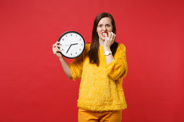 Nervous young woman in yellow fur sweater gnawing nails, holding round clock isolated on red...
