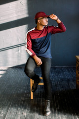 Portrait of an African-American hipster guy dressed in a red fleece sweater and red cap at the studio. Isolated on dark textured background sitting in a studio on a wooden chair