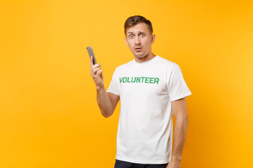 Young man portrait in white t-shirt with written inscription green title volunteer talking on mobile phone isolated on yellow background. Voluntary free assistance help, charity grace work concept.