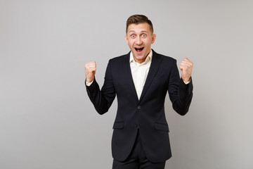 Surprised young business man in classic black suit, shirt doing winner gesture keeping mouth wide open isolated on grey wall background. Achievement career wealth business concept. Mock up copy space.