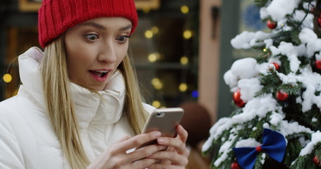 Blonde beautiful young woman holding her smartphone in hands, looking at the screen and making surprising face. Christmas tree and decorated street behind her. Outside. Close up. Portrait shot.