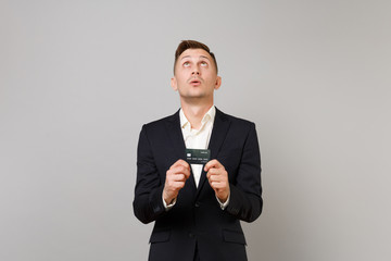 Portrait of young business man in classic black suit, shirt looking up, holding credit bank card isolated on grey background in studio. Achievement career wealth business concept. Mock up copy space.