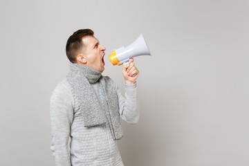 Side view of young man in gray sweater, scarf scream in megaphone isolated on grey wall background in studio. Healthy fashion lifestyle people sincere emotions cold season concept. Mock up copy space.