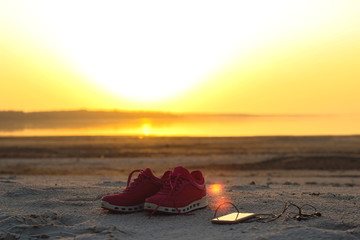 Sports set. Pink sneakers and a mobile phone with headphones close-up on a sandy beach by the sea during sunset..